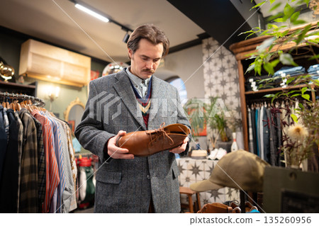 Serious handsome man customer thinking on buying fancy leather shoes in second hand vintage shop. Serious handsome man customer thinking on buying fancy leather shoes in second hand vintage shop. 135260956