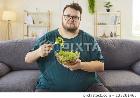 Unhappy man eating salad on sofa at home, struggling with healthy choices Unhappy man eating salad on sofa at home, struggling with healthy choices 135261465