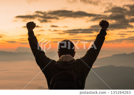 Silhouette of a hiker raising fists at sunrise above layered mountains and warm clouds. Victory, freedom and motivation concept, scenic travel background with copy space for headlines. Silhouette of a hiker raising fists at sunrise above layered mountains and warm clouds. Victory, freedom and motivation concept, scenic travel background with copy space for headlines. 135264263