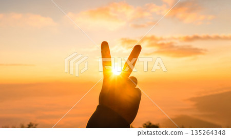 Silhouette hand making peace sign framing the sun at golden sunrise, warm orange sky and soft clouds. Minimal travel background with copy space for victory, freedom and inspiration. Silhouette hand making peace sign framing the sun at golden sunrise, warm orange sky and soft clouds. Minimal travel background with copy space for victory, freedom and inspiration. 135264348