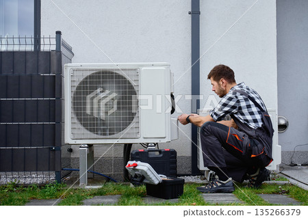 Technician servicing outdoor air source heat pump unit near house wall 135266379