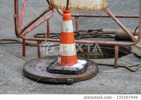 A rusty orange and white traffic cone is on the ground next to a metal grate A rusty orange and white traffic cone is on the ground next to a metal grate 135267449