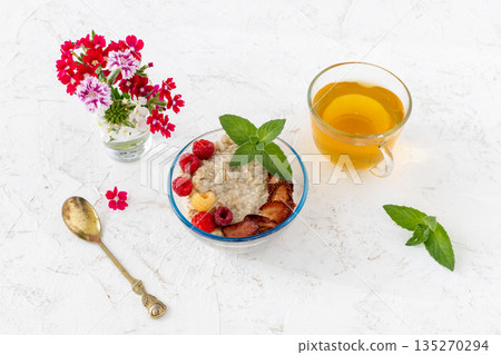 Oatmeal porridge with berries in a glass bowl and a cup of tea on the white table. Oatmeal porridge with berries in a glass bowl and a cup of tea on the white table. 135270294