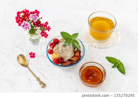Oatmeal porridge with berries in a glass bowl and a cup of tea on the white table. 135270295