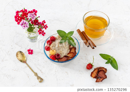 Oatmeal porridge with berries in a glass bowl and a cup of tea on the white table. 135270296