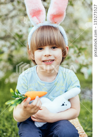 Easter Egg Hunt with Child Carrying Bright Plastic Eggs. Smiling child wearing bunny ears holding a wicker basket filled with colorful Easter eggs in a blooming spring garden Easter Egg Hunt with Child Carrying Bright Plastic Eggs. Smiling child wearing bunny ears holding a wicker basket filled with colorful Easter eggs in a blooming spring garden 135273527