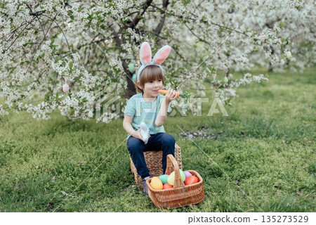 Easter Egg Hunt with Child Carrying Bright Plastic Eggs. Smiling child wearing bunny ears holding a wicker basket filled with colorful Easter eggs in a blooming spring garden 135273529