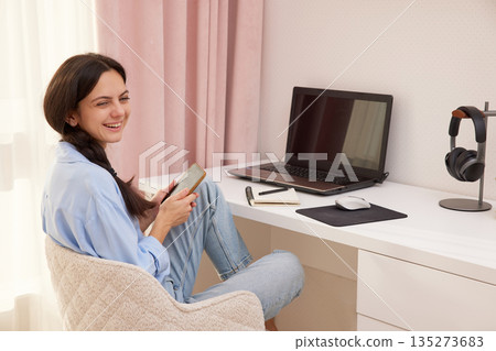 Happy woman holding a smartphone, sitting at a desk with a laptop 135273683