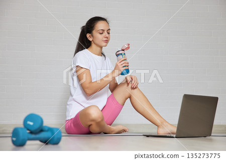 Young woman hydrating with a water bottle after exercising, using a laptop for online fitness 135273775