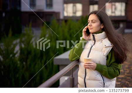 Woman standing outdoors, talking on smartphone and holding a takeaway coffee cup Woman standing outdoors, talking on smartphone and holding a takeaway coffee cup 135273799