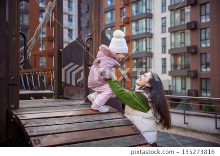 Happy mother raising up daughter on playground with city buildings in background 135273816