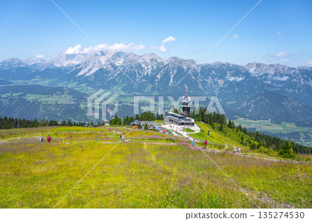 Breathtaking landscape showcases the Dachstein mountain range from Planai. Visitors explore lush green meadows with the summit station in the foreground, under a clear blue sky. Breathtaking landscape showcases the Dachstein mountain range from Planai. Visitors explore lush green meadows with the summit station in the foreground, under a clear blue sky. 135274530