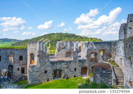 Visitors stroll through the courtyard of Rabi Castle, surrounded by ancient stone walls and lush green hills under a bright blue sky. The historic site offers a glimpse into the past. 135274594