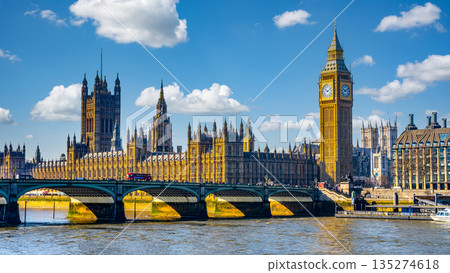 Houses of Parliament and Big Ben rise majestically against a clear blue sky. The River Thames flows calmly beneath Westminster Bridge as a red bus crosses, showcasing London vibrant atmosphere. 135274618
