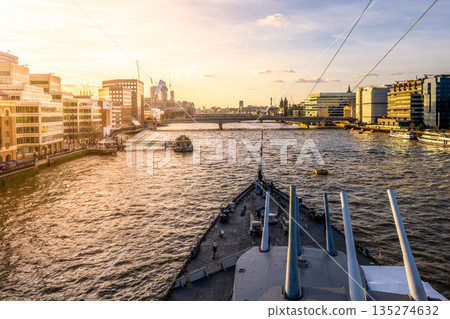 Turrets stand tall on HMS Belfast as the sun sets behind the London skyline. The River Thames flows nearby, with city life bustling along the waterfront. 135274632