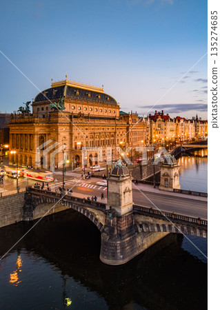 The aerial scene shows the National Theatre in Prague illuminated at dusk. The Smetana embankment and Rasin bridge are visible, creating a lively atmosphere in Czechia. The aerial scene shows the National Theatre in Prague illuminated at dusk. The Smetana embankment and Rasin bridge are visible, creating a lively atmosphere in Czechia. 135274685