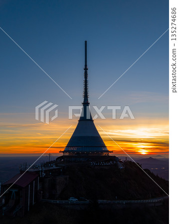 The aerial view shows the Jested mountain and hotel at sunset. The sky displays shades of color as the sun sets behind the iconic structure. This moment captures evening at Liberec. The aerial view shows the Jested mountain and hotel at sunset. The sky displays shades of color as the sun sets behind the iconic structure. This moment captures evening at Liberec. 135274686