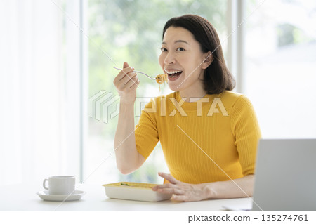 一張五十多歲女性在辦公桌前邊工作邊吃午餐的照片 一張五十多歲女性在辦公桌前邊工作邊吃午餐的照片 135274761