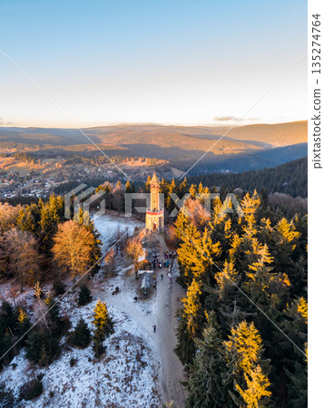 People gather near Stepanka lookout tower in the Jizera Mountains during sunset. The tower is surrounded by trees and snow, creating a beautiful winter scene. 135274764