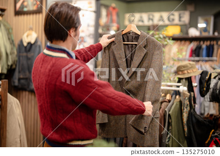 Stylish man buying trendy fashionable items in vintage clothing store, holding blazer on hanger. 135275010