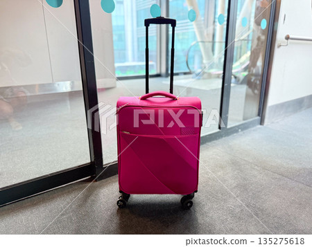 Pink carry on suitcase near glass airport entrance. Travel preparation, personal mobility, and departure mindset through luggage and terminal space Pink carry on suitcase near glass airport entrance. Travel preparation, personal mobility, and departure mindset through luggage and terminal space 135275618