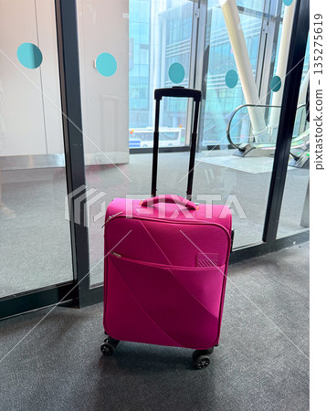 Pink carry on suitcase standing near airport glass doors. Travel departure, mobility, and modern transport through personal luggage and terminal architecture Pink carry on suitcase standing near airport glass doors. Travel departure, mobility, and modern transport through personal luggage and terminal architecture 135275619