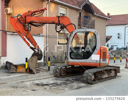 Excavator working on residential street beside suburban houses at sunset. Road repair, urban infrastructure, and neighborhood development through construction activity and municipal service Excavator working on residential street beside suburban houses at sunset. Road repair, urban infrastructure, and neighborhood development through construction activity and municipal service 135275620