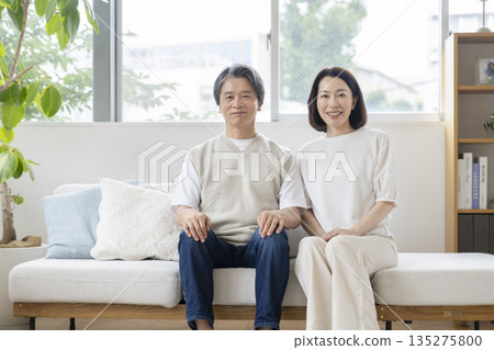 一對五十多歲的夫婦並排坐在沙發上,對著鏡頭微笑。 一對五十多歲的夫婦並排坐在沙發上,對著鏡頭微笑。 135275800
