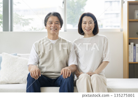 Image of a couple in their 50s sitting side by side on the sofa and smiling at the camera 135275801