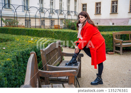 Woman walking in park in Prague during winter without snow wearing red coat. Calm urban walking, everyday lifestyle and seasonal city routine in real life European environment 135276381