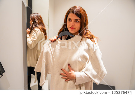 Woman trying on clothes in fitting room while standing in front of mirror inside fashion store, Shopping experience, personal style choice and consumer decision process in real life retail environment Woman trying on clothes in fitting room while standing in front of mirror inside fashion store, Shopping experience, personal style choice and consumer decision process in real life retail environment 135276474