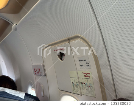 Emergency exit door and safety instructions inside aircraft cabin. Passenger security, aviation regulation, and flight preparedness through signage, procedure, and design 135280023