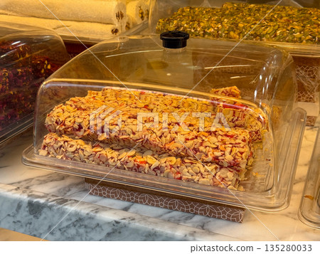 Layered almond pastry dessert displayed under glass dome in Istanbul confectionery. Traditional Turkish sweets, nut pastry, and bakery culture through artisanal food and local gastronomy Layered almond pastry dessert displayed under glass dome in Istanbul confectionery. Traditional Turkish sweets, nut pastry, and bakery culture through artisanal food and local gastronomy 135280033