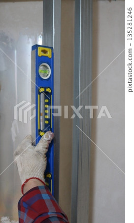 A gloved hand vertically holds a blue level on a metal profile in a room structure and measures the angle of inclination, taking measurements during the construction of an indoor partition 135281246