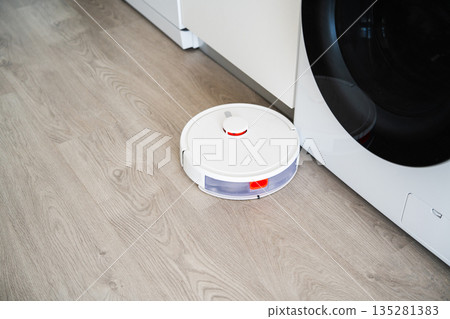 Robot vacuum cleaner cleaning floor next to washing machine, smart home automation and modern household cleaning technology 135281383