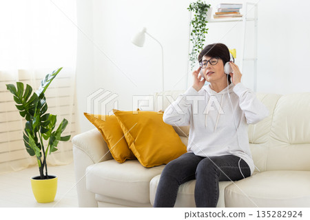 Woman listening to music with headphones on sofa in modern living room. Audio relaxation, digital entertainment, and mindful leisure through home technology. 135282924