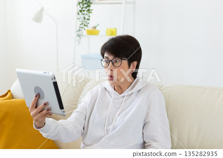 Woman making videocall on tablet while sitting on sofa in modern apartment. Online communication, remote meeting, and digital interaction in home interior. 135282935