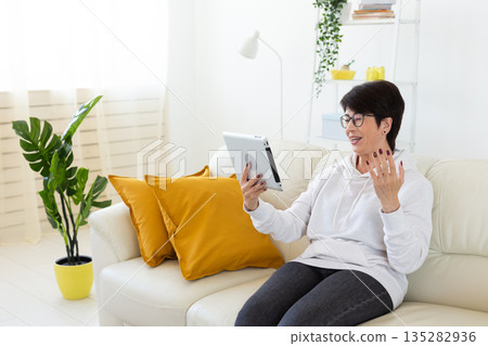 Woman making videocall on tablet while sitting on sofa in modern apartment. Online communication, remote meeting, and digital interaction in home interior. 135282936
