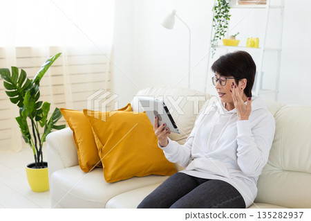 Woman making videocall on tablet while sitting on sofa in modern apartment. Online communication, remote meeting, and digital interaction in home interior. 135282937