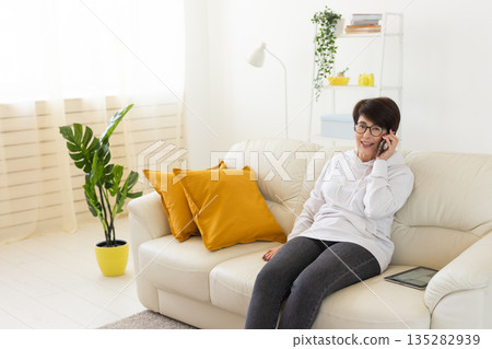 Woman talking on smartphone on white sofa in modern living room. Home communication, remote conversation, and digital lifestyle inside bright apartment interior. Woman talking on smartphone on white sofa in modern living room. Home communication, remote conversation, and digital lifestyle inside bright apartment interior. 135282939