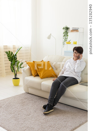 Woman talking on smartphone on white sofa in modern living room. Home communication, remote conversation, and digital lifestyle inside bright apartment interior. 135282940