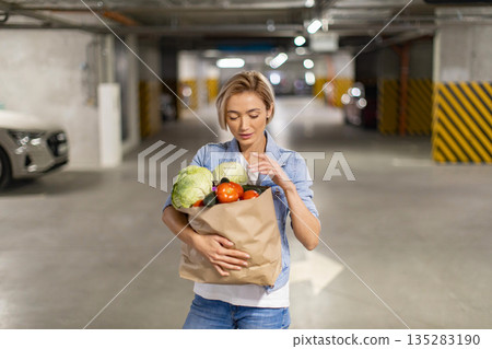 A woman carries a bag of fresh produce in an underground parking garage, likely after a shopping trip A woman carries a bag of fresh produce in an underground parking garage, likely after a shopping trip 135283190