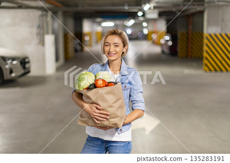 A smiling woman carries a bag of fresh produce in an underground parking garage after shopping 135283191