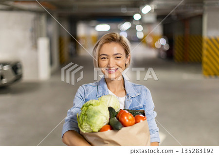 A smiling woman carries a bag of fresh produce in an underground parking garage, ready to prepare a healthy meal A smiling woman carries a bag of fresh produce in an underground parking garage, ready to prepare a healthy meal 135283192