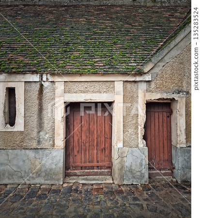 Rustic building with moss covered tiled roof and textured walls. Two old wooden doors with vertical planks next to a narrow window. Cobblestone street pavement Rustic building with moss covered tiled roof and textured walls. Two old wooden doors with vertical planks next to a narrow window. Cobblestone street pavement 135283524