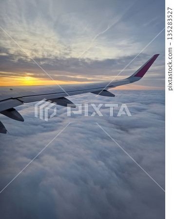 Peaceful aerial view from airplane window to the large wing soaring over a sea of clouds with the sun rising above the horizon, casting a warm glow and illuminating the cloudscape during flight 135283527