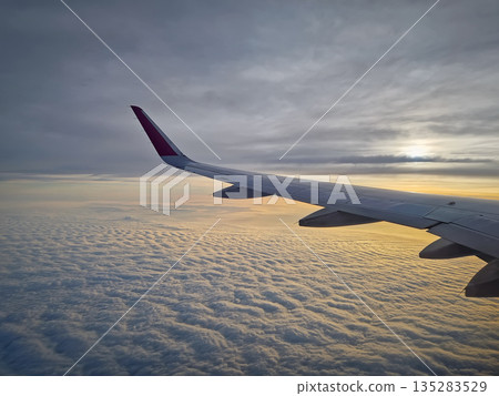 Peaceful aerial view from airplane window to the large wing soaring over a sea of clouds with the sun rising above the horizon, casting a warm glow and illuminating the cloudscape during flight Peaceful aerial view from airplane window to the large wing soaring over a sea of clouds with the sun rising above the horizon, casting a warm glow and illuminating the cloudscape during flight 135283529