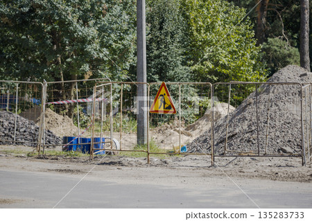 Workers are preparing a construction area marked by a fence and a warning sign. Dirt and materials are visible in the background among trees 135283733
