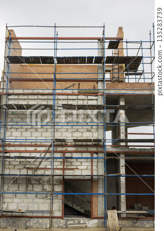 Workers are constructing a building with scaffolding around it. The site shows walls made of bricks and concrete, indicating ongoing work Workers are constructing a building with scaffolding around it. The site shows walls made of bricks and concrete, indicating ongoing work 135283739