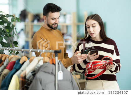 Woman selects handbag and checks its quality using QR code. Man helps her Woman selects handbag and checks its quality using QR code. Man helps her 135284107